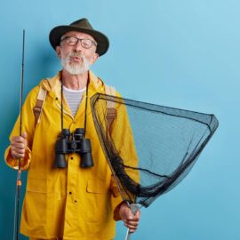 man standing against blue background with fishing equipment, rain coat, and binoculars.